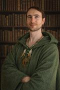 Portrait d'Iphégore Ossenoire souriant, en tenue de chamane, devant une bibliothèque de livres anciens